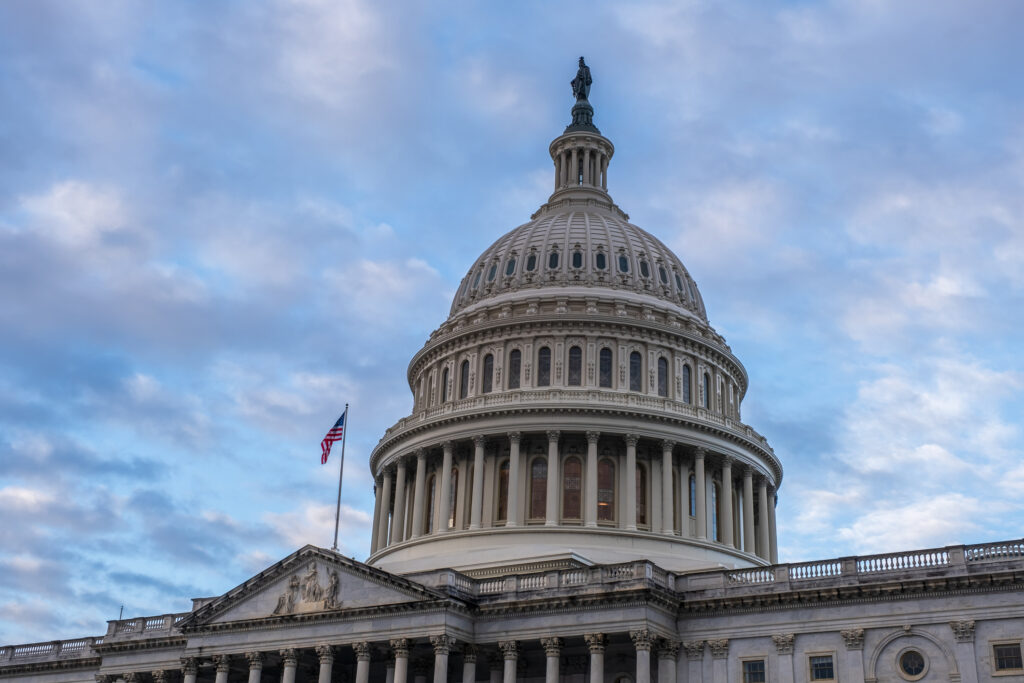 The dome of the United States Capitol building with an American flag waving beside it, set against a partly cloudy blue sky.