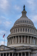 The dome of the United States Capitol building with an American flag waving beside it, set against a partly cloudy blue sky.