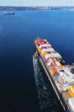 Aerial view of a container ship nearing port with the assistance of a tugboat.